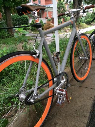 A single-speed bike with orange rims and a belt drive in place of a chain; a simple, clean design that requires less maintenance while the sleek lines make it easy to modify and customize, and without all the added gears, this is a cheaper to purchase.
