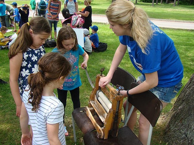 A high school student volunteer helps children take turns using an antique laundry machine to compare cleaning methods from the 1800s with modern ways
