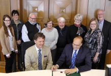 Peterborough Symphony Orchestra Music Director and Conductor Michael Newnham sits beside Mayor Daryl Bennett as he signs the proclamation of Symphony Week in Peterborough, with staff and supporters of the Peterborough Symphony Orchestra in the background (photo: Bruce Head / kawarthaNOW)