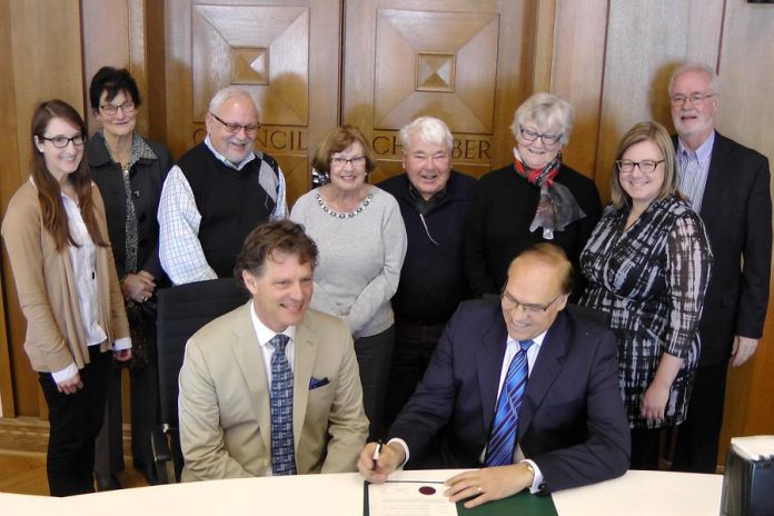 Peterborough Symphony Orchestra Music Director and Conductor Michael Newnham sits beside Mayor Daryl Bennett as he signs the proclamation of Symphony Week in Peterborough, with staff and supporters of the Peterborough Symphony Orchestra in the background (photo: Bruce Head / kawarthaNOW)