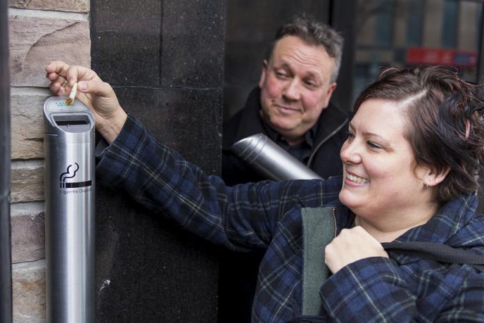 Volunteer Donna Reid, who also coordinates the annual Butt Blitz in Peterborough, disposes of a cigarette butt in one of the new receptacles in downtown Peterborough as DBIA Executive Director Terry Guiel looks on. The DBIA is offering the free receptacles to its members. (Photo: Peterborough DBIA)