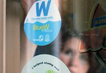 GreenUP Water Programs Coordinator Jenn McCallum places a BlueW Ptbo decal in the window at Black Honey Café on Hunter Street in downtown Peterborough. Look for the decal across the Kawarthas at businesses and public service buildings, marking them as destinations for re-filling up your reusable water bottle for free, or check bluewptbo.ca for a full listing. (Photo: Karen Halley / GreenUP)