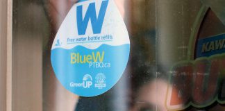 GreenUP Water Programs Coordinator Jenn McCallum places a BlueW Ptbo decal in the window at Black Honey Café on Hunter Street in downtown Peterborough. Look for the decal across the Kawarthas at businesses and public service buildings, marking them as destinations for re-filling up your reusable water bottle for free, or check bluewptbo.ca for a full listing. (Photo: Karen Halley / GreenUP)