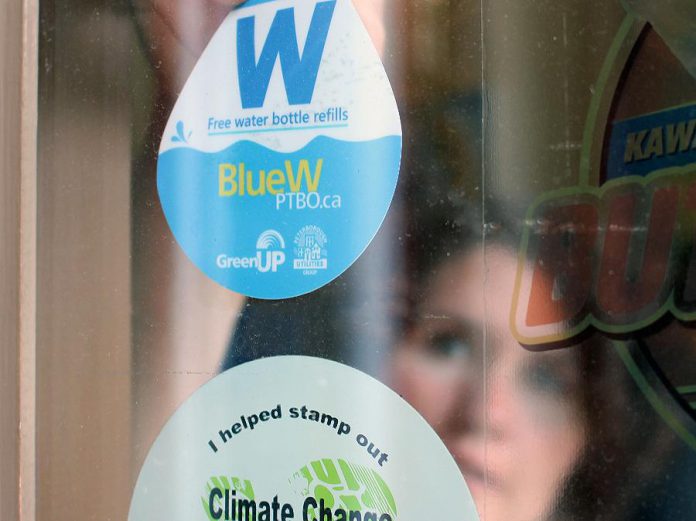 GreenUP Water Programs Coordinator Jenn McCallum places a BlueW Ptbo decal in the window at Black Honey Café on Hunter Street in downtown Peterborough. Look for the decal across the Kawarthas at businesses and public service buildings, marking them as destinations for re-filling up your reusable water bottle for free, or check bluewptbo.ca for a full listing. (Photo: Karen Halley / GreenUP)