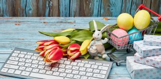 Colored Easter eggs in a miniature shopping cart, gift boxes, tulips and computer keyboard and mouse on a wooden background.