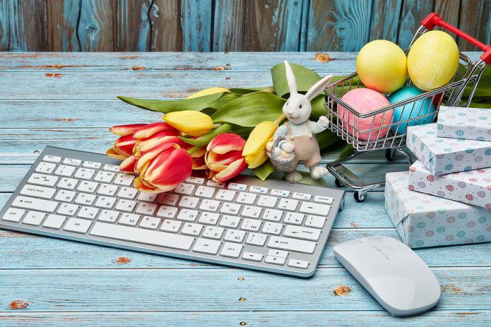 Colored Easter eggs in a miniature shopping cart, gift boxes, tulips and computer keyboard and mouse on a wooden background.