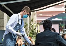 A waiter with a mask serves two customers on a patio