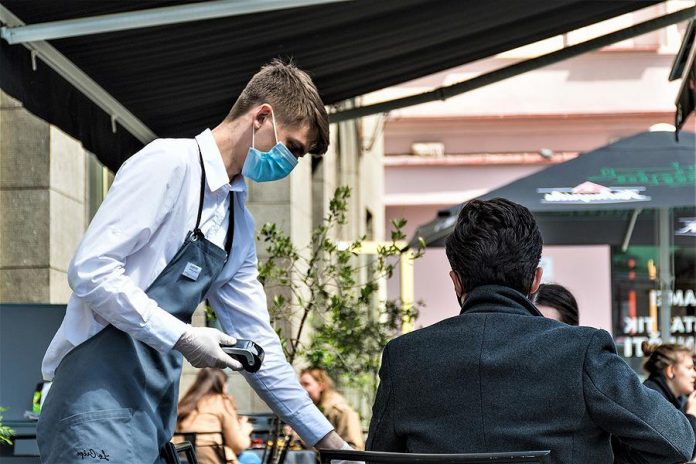 A waiter with a mask serves two customers on a patio