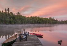 Muskoka chairs on a dock with canoes at a lake. (Stock photo)