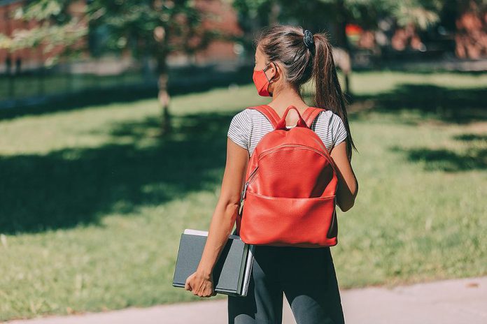 A young masked female student wearing a backpack and carrying books. (Stock photo)