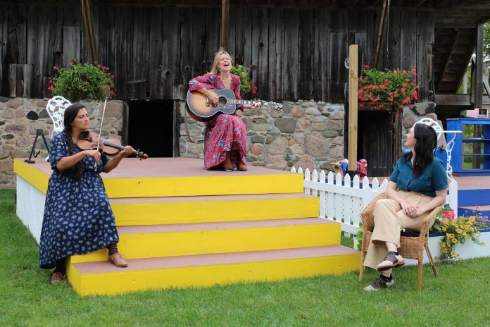 Singer-songwriter Kate Suhr belts out a tune as musician Saskia Tomkins provides accompaniment and storyteller Megan Murphy looks on during a media day at 4th Line Theatre on Millbrook on August 12, 2021, promoting The Verandah Society In Residence. Created and performed Murphy and Suhr, the show is a hybrid of storytelling and music and runs for two weeks in August at the Winslow Farm. (Photo: Hannah Abrahamse / kawarthaNOW)