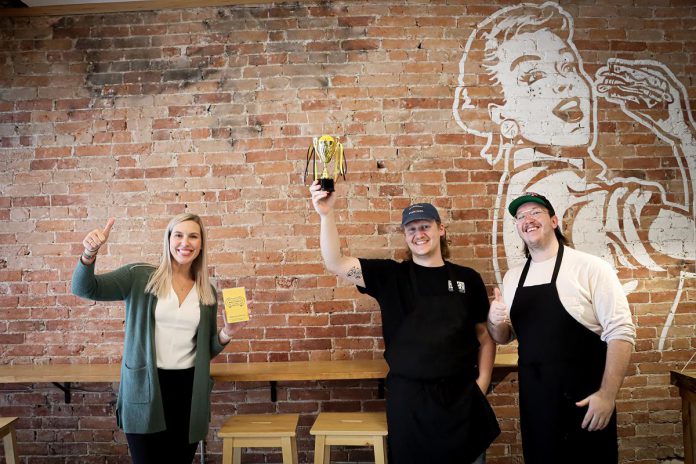  Kawartha Food Share general manager Ashlee Aitken (left) celebrating the Peterborough Mac + Cheese Festival champion with Sam's Place staff members Eddy Sweeney (holding the "cheesy" trophy) and Owen Walsh.  As part of Sam's Place's win, the Peterborough Downtown Business Improvement Area made a $500 donation to Kawartha Food Share in the restaurant's name. (Photo courtesy of Peterborough DBIA)