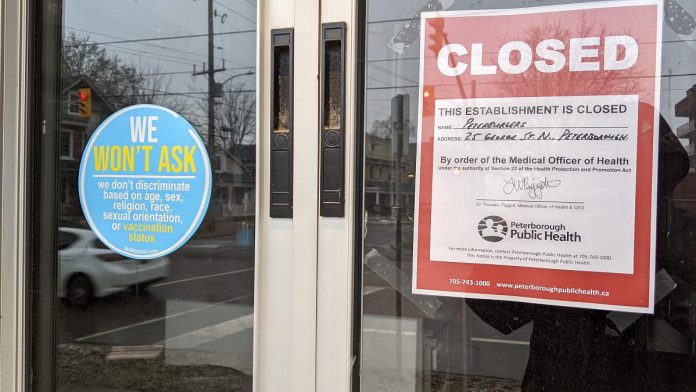 A notice in the window of Peterburgers at 25 George Street North in Peterborough that the restaurant has been closed by order of the medical officer of health, beside a decal posted by the restaurant stating that it does not ask for proof of vaccination, a requirement under the province's Reopening Ontario Act. (Photo: kawarthaNOW)