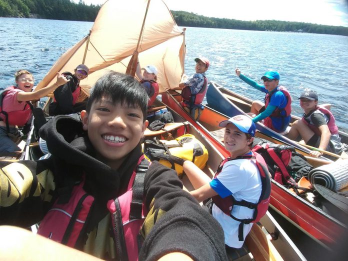 A group of international students take a short break from paddling to pose for a selfie while the wind does the work of propelling their canoe raft along. The Learning Languages in the Outdoors program provides students with the opportunity to expand their English languages skills while experiencing Canadian culture.  (Photo courtesy of The Land Canadian Adventures)