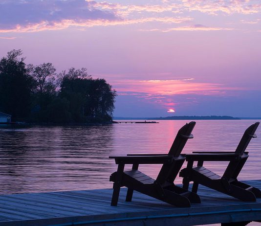 Two empty Muskoka chairs on a dock on a lake at sunset. (Stock photo)