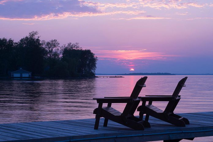 Two empty Muskoka chairs on a dock on a lake at sunset. (Stock photo)