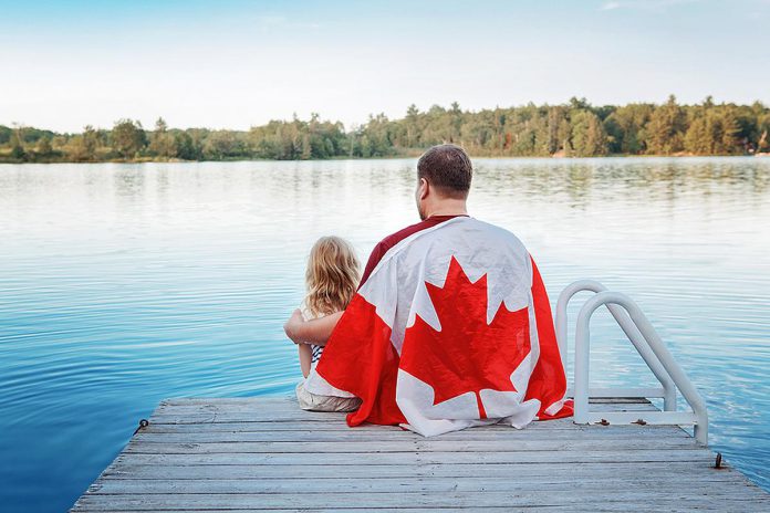 A man wrapped in the Canadian flag sitting on a dock with his young daughter. (Stock photo)