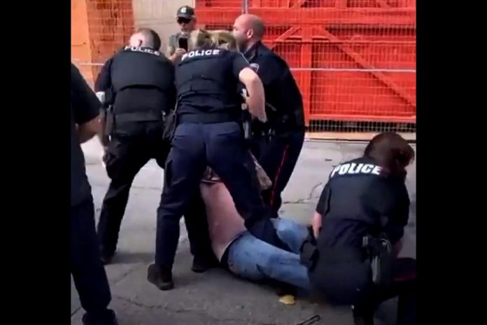 Peterborough police officers arrest Frank Curtin during a protest at the Peterborough police station on August 13, 2022. A follower of self-proclaimed "Queen of Canada" Romana Didulo, Curtin organized and led the protest to "arrest" Peterborough police officers after a call from Didulo, who espouses a mix of sovereign citizen beliefs, QAnon conspiracy theories, and new age spirituality. Didulo also claims to be an extraterrestrial being with supernatural powers. (kawarthaNOW screenshot of Twitter video)