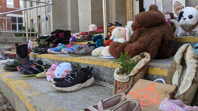 Children's shoes, toys, and offerings on the steps of Peterborough City Hall in June 2021, part of a community memorial created in response to the discovery of the remains of 215 Indigenous children buried at the former Kamloops Indian Residential School in British Columbia. As well as remembering and honouring those lost to residential schools and the thousands of survivors, National Day for Truth and Reconciliation is a day for Canadians to educate themselves about the heritage, culture, stories, and experiences of First Nations, Inuit, and Métis peoples. (Photo: Bruce Head / kawarthaNOW)