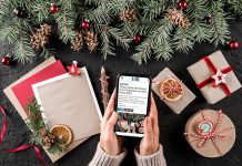 A woman sitting at a Christmas-decorated table holding a mobile phone displaying kawarthaNOW's Christmas and New Year's holiday hours story. (kawarthaNOW photo)
