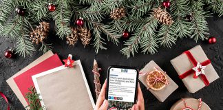 A woman sitting at a Christmas-decorated table holding a mobile phone displaying kawarthaNOW's Christmas and New Year's holiday hours story. (kawarthaNOW photo)