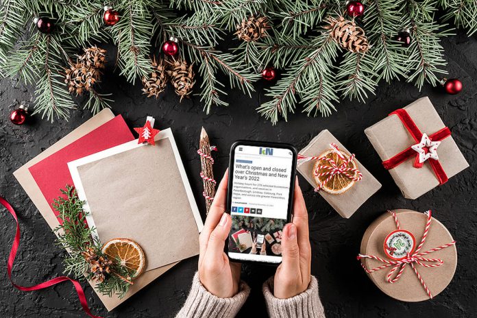 A woman sitting at a Christmas-decorated table holding a mobile phone displaying kawarthaNOW's Christmas and New Year's holiday hours story. (kawarthaNOW photo)