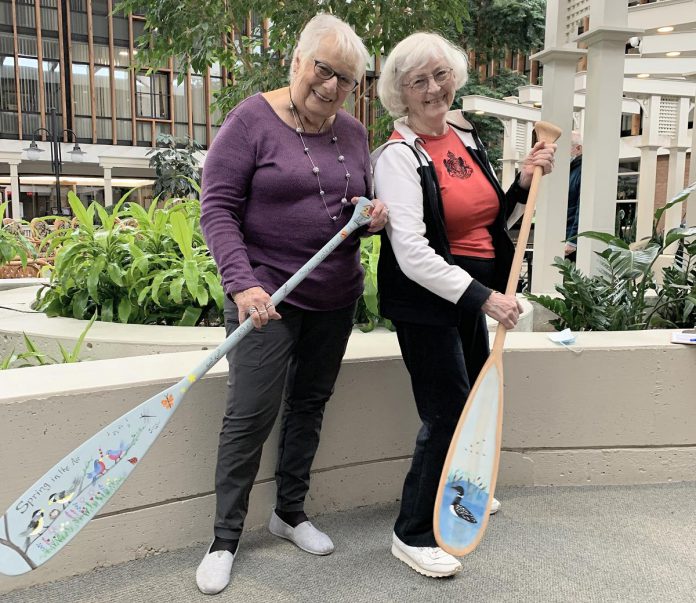 Two residents of The Gardens of Peterborough holding their painted paddles: "Spring in the Air" (left) and "Call of the Loon". These and the other 29 paddles in the exhibit will be auctioned off during March 2023 with proceeds supporting the Downtown Green Team, a DBIA partnership with the One City Employment Program  a seasonal gardening team to employ people experiencing barriers to traditional employment. (Photo courtesy of Peterborough DBIA)