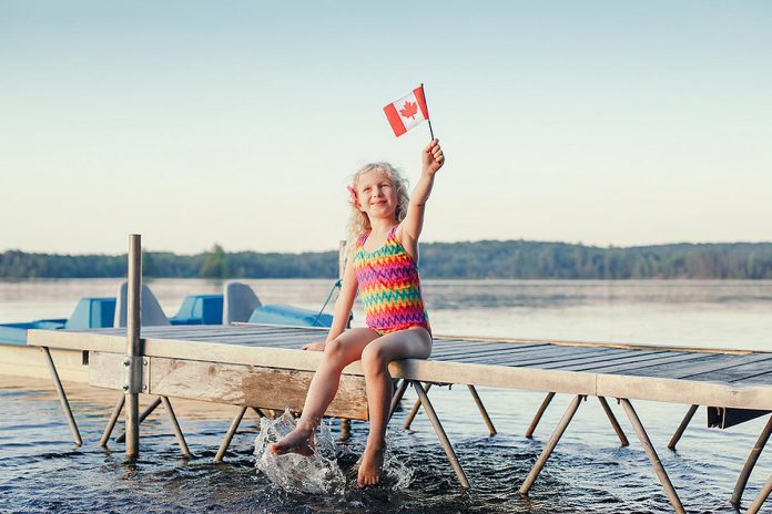 A young girl sitting on a dock and waving a small Canadian flag. (Stock photo)