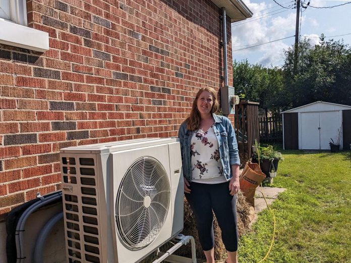 Peterborough homeowner Deanna VandenBroek stands next to her new electric heat pump. VandenBroek chose heat pump technology with an electric furnace as a secondary heat source. (Photo: Clara Blakelock / GreenUP)