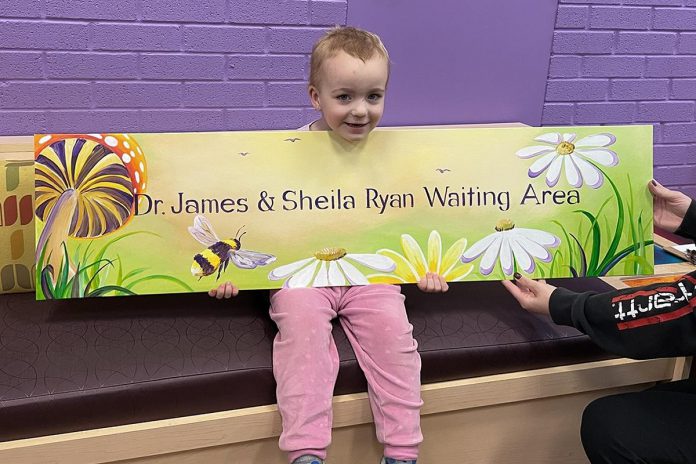 Maisie, one of the 6,200 children and youth served by Five Counties Children's Centre in the past year, helps display a sign the charitable organization serving children with special needs in the greater Kawarthas region will hang in the waiting area of its Peterborough location in recognition of a $227,000 donation from retired Peterborough couple James and Sheila Ryan to help reduce wait times. (Photo courtesy of Five Counties Children's Centre)