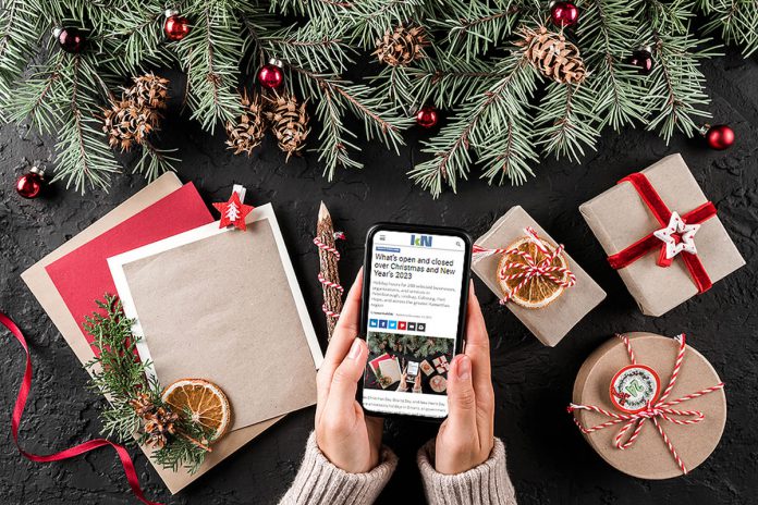 Hands holding a smartphone displaying kawarthaNOW's 2023 holiday hours listing with a Christmas scene in the background. (Photo: kawarthaNOW)