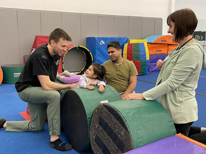 Kawartha Gymnastics coach Nathan (left) gets a smile from two-year-old Mirha as she takes part in a new adaptive gymnastics program at the club. Looking on are Mirha's dad Mansoor and Five Counties Children's Centre recreation therapist Colleen Ristok. (Photo: Five Counties Children's Centre)