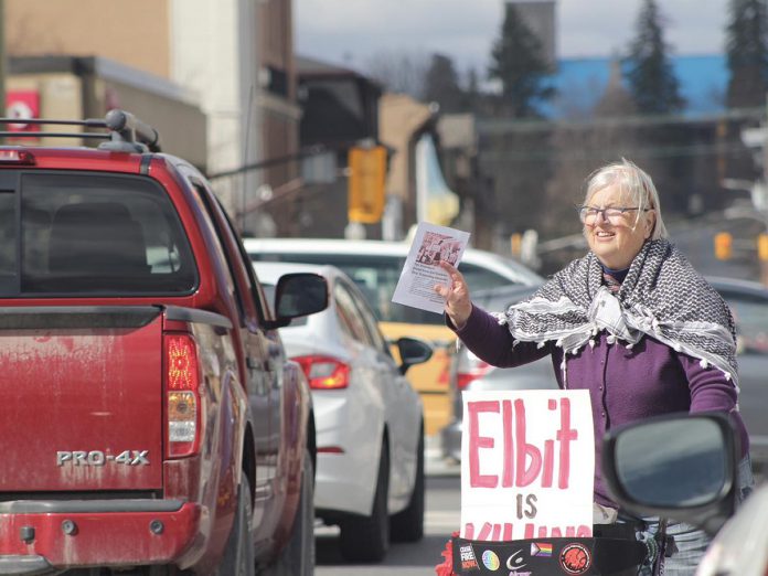 A protester offers a leaflet to passing vehicles at the Scotiabank branch at Hunter and Water streets in downtown Peterborough on March 15, 2024. The protest was one of dozens at Scotiabank branches across the country demanding the bank divest its remaining holdings in controversial Israeli  weapons manufacturer Elbit Systems Ltd. (Photo: Nogojiwanong 2 Palestine)