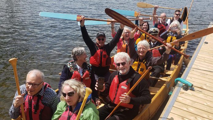 Lieutenant Governor of Ontario Edith Dumont (holding her paddle in the air, third from bottom on the right) was among the dignitaries who paddled one of the Canadian Canoe Museum's Voyageur canoes into Little Lake during a grand opening celebration on May 11, 2024 for the now-completed new museum at 2077 Ashburnham Drive. (Photo: Jeannine Taylor / kawarthaNOW)