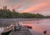 Two Muskoka chairs on a dock at a lake with a canoe and kayak nearby. (Stock photo)