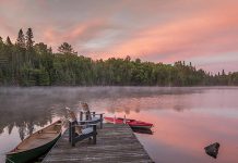 Two Muskoka chairs on a dock at a lake with a canoe and kayak nearby. (Stock photo)