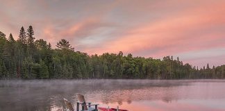 Two Muskoka chairs on a dock at a lake with a canoe and kayak nearby. (Stock photo)