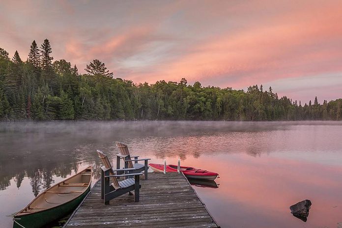 Two Muskoka chairs on a dock at a lake with a canoe and kayak nearby. (Stock photo)