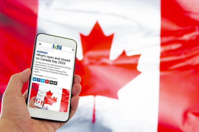 A hand holding a mobile phone showing kawarthaNOW's Canada Day hours story, with a Canadian flag in the background. (Photo: kawarthaNOW)