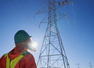 Hydro One worker looking up at other workers on a transmission tower. (Photo: Hydro One)