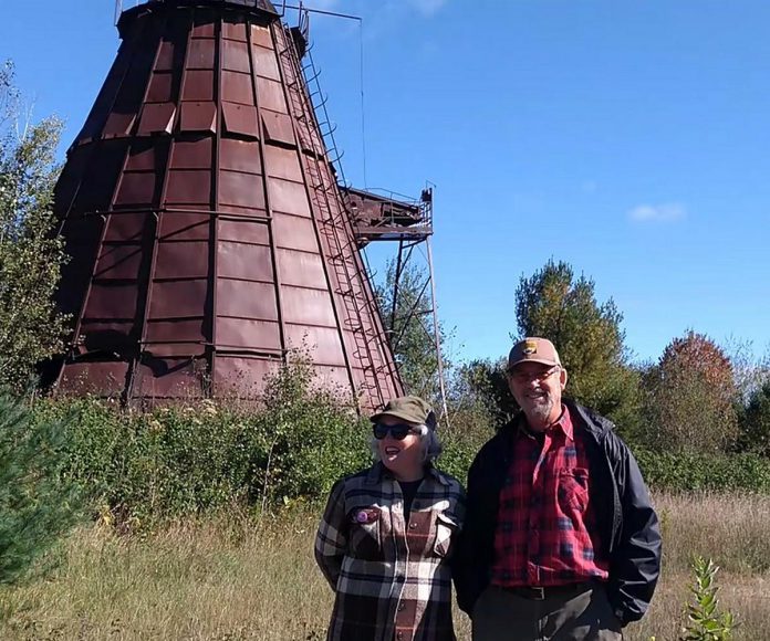 Highlands East artists Victoria Ward and Gary Blundell at the sawdust burner that inspired the artwork featured in their "Burner Herzog" exhibit on display from August 22 to November 23, 2024 at the Agnes Jamieson Gallery at the Minden Hills Cultural Centre. The married artists first stumbled upon the site of the lumber mill near their log home 15 years ago and have since revisited the location many times, watching nature take the land back from the abandoned site. Along with the exhibit, there will be a trip to visit the sawdust burner as well as a film screening. (Photo courtesy of Victoria Ward and Gary Blundell)