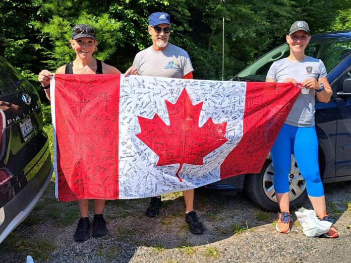 Canal Pursuit For Mental Health founder Clay Williams (middle) in 2023 with the Canadian flag that serves as a baton for the annual relay run. During the run, he's asked people along the route to sign the flag if they or someone they know has mental health issues. (Photo courtesy of Clay Williams)