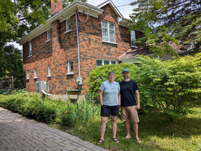 Madeleine Endicott and Steve McMurtry in front of their century home near Bonnerworth Park in Peterborough. Because of the brick exterior, cellulose insulation was installed through holes in the interior walls of the home. The couple's air source heat pump with electric backup, which replaced their natural gas furnace, can be seen along the side of the home. (Photo: Clara Blakelock / GreenUP)