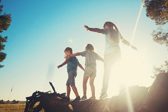 Three children outside in the warm weather balancing on a fallen tree near a field. (Stock photo)