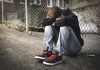 A teenage boy sitting on the street against a fence with his head down on crossed arms on his knees. (Stock photo)