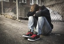 A teenage boy sitting on the street against a fence with his head down on crossed arms on his knees. (Stock photo)