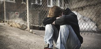 A teenage boy sitting on the street against a fence with his head down on crossed arms on his knees. (Stock photo)