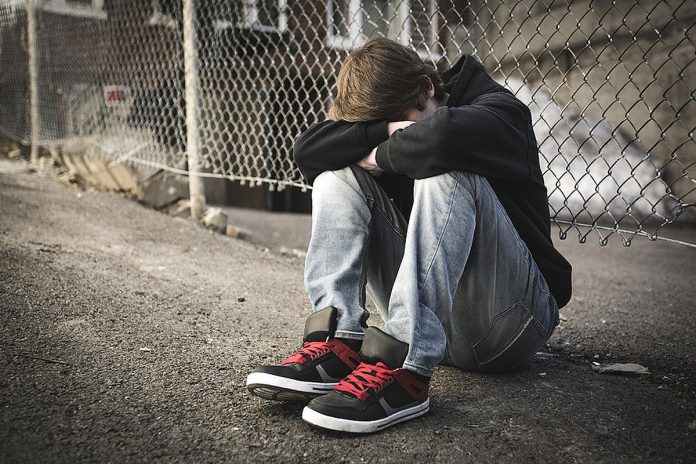 A teenage boy sitting on the street against a fence with his head down on crossed arms on his knees. (Stock photo)