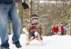 A outdoors winter scene showing a man pulling a young boy on a sled through the woods with a young girl and boy following in the background. (Stock photo)