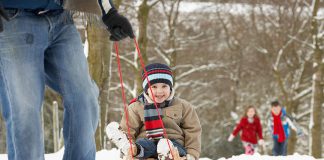 A outdoors winter scene showing a man pulling a young boy on a sled through the woods with a young girl and boy following in the background. (Stock photo)